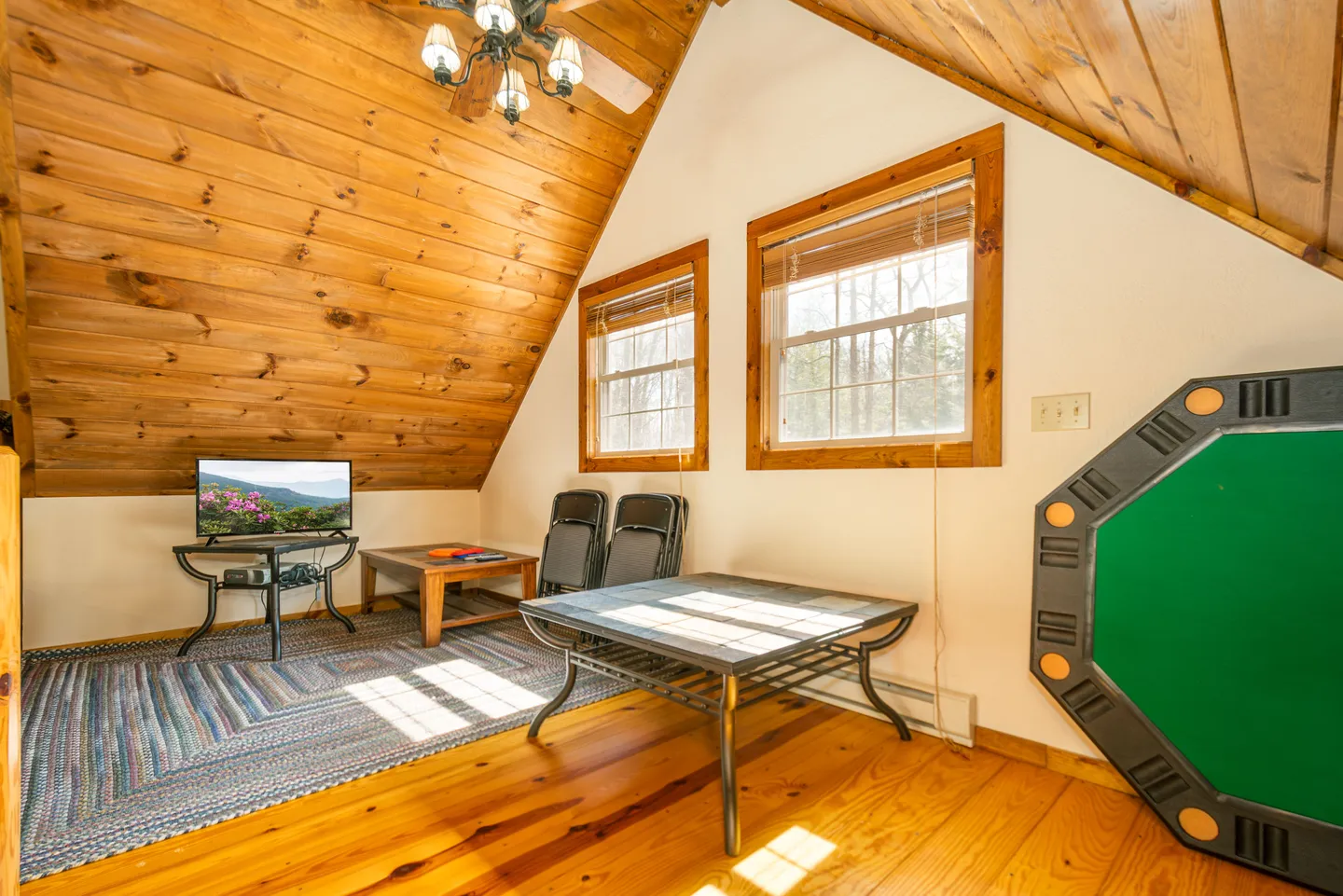 Loft or attic room with wood-paneled angled ceilings, a small TV on a stand, folding chairs, small tables, and a poker tabletop leaned against the wall.