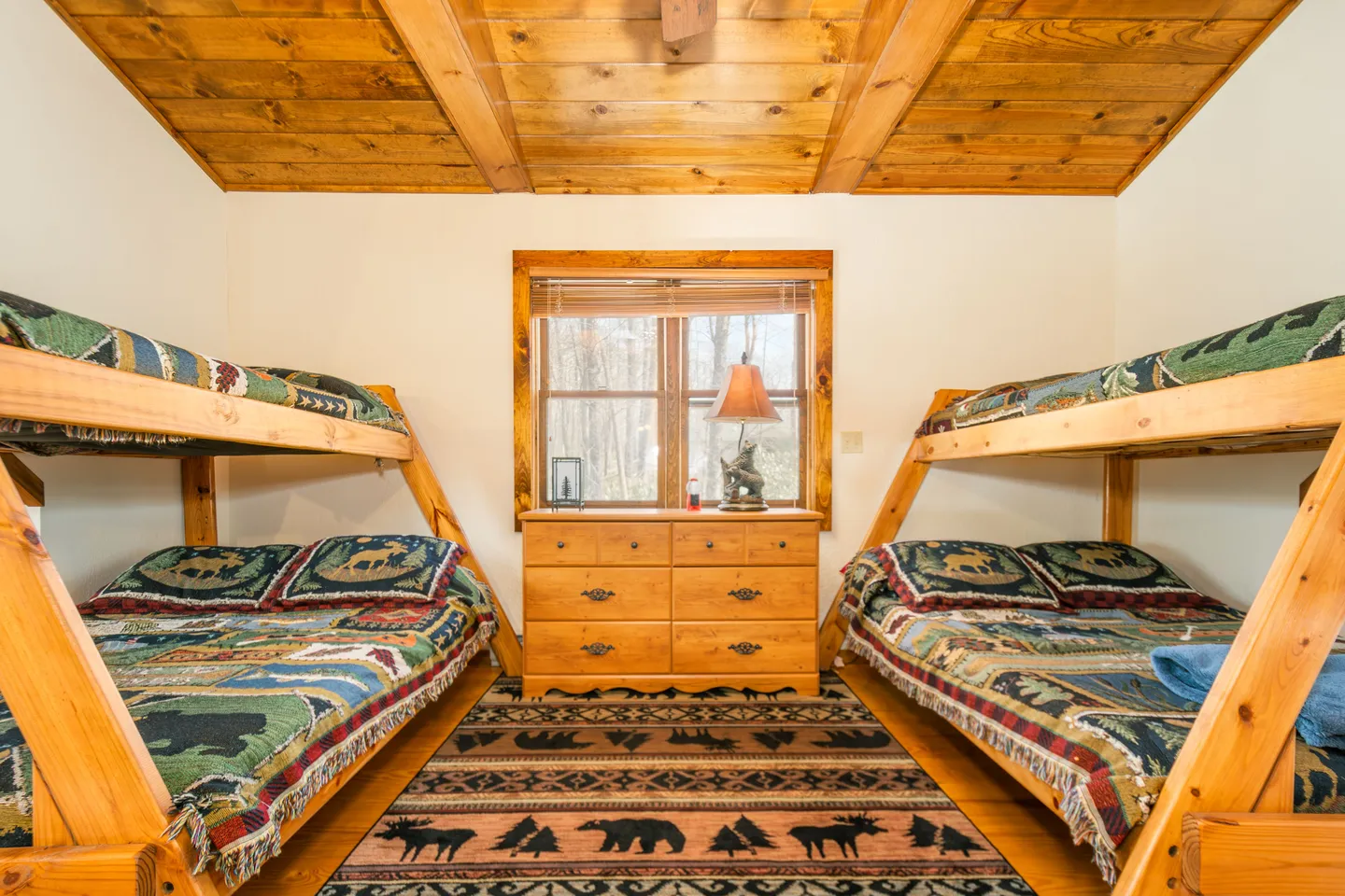 Bedroom with two wooden bunk beds, a central wooden dresser under a window, rustic patterned bedding, and a bear and moose motif rug.