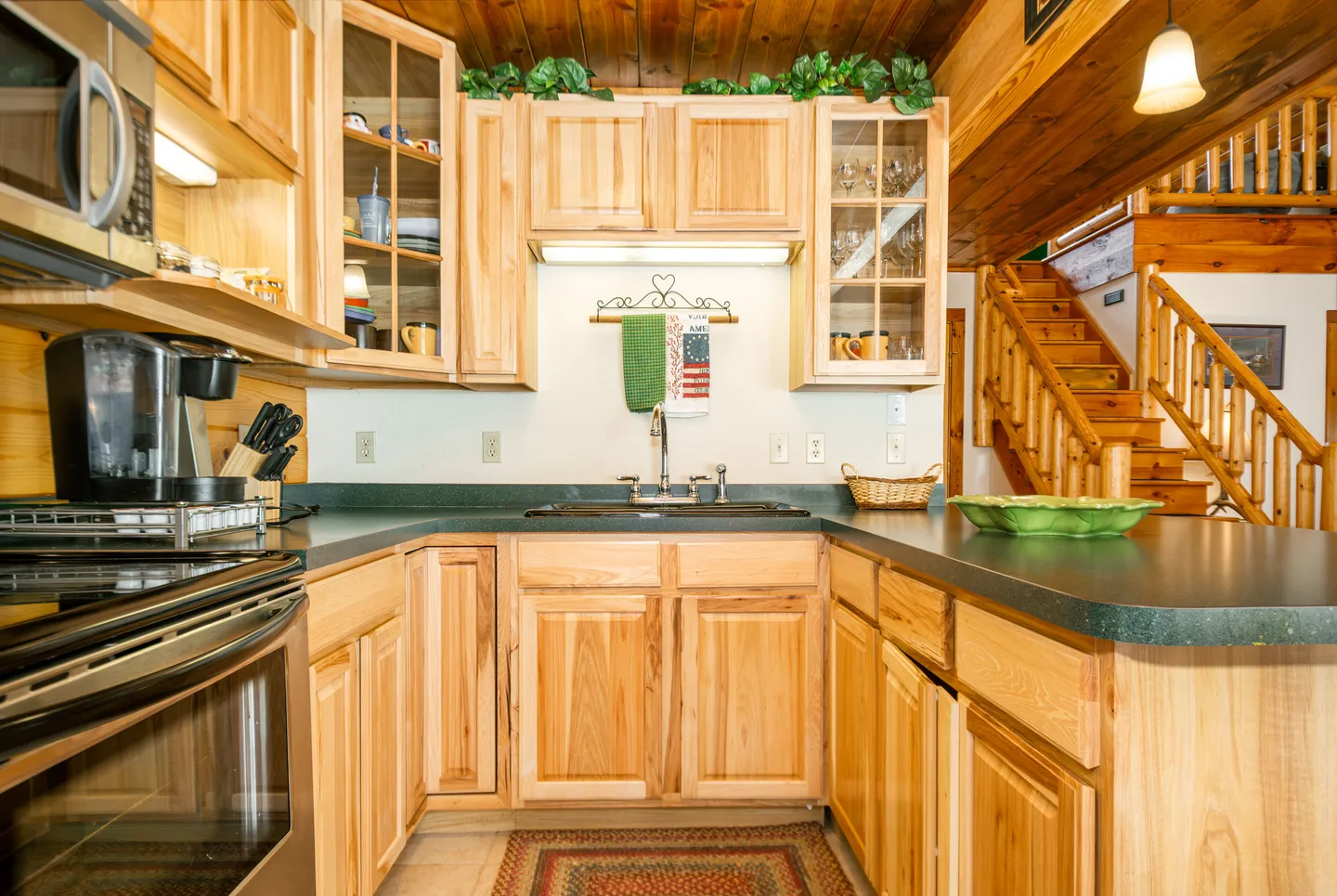Kitchen in a log cabin with natural wood cabinets, dark countertops, sink, and view of a wooden staircase in the background.
