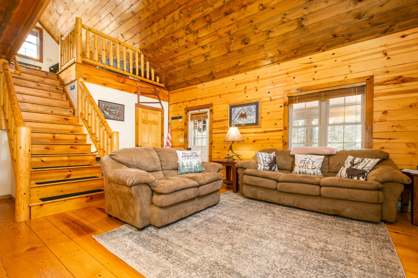 Living room with wood-paneled walls and ceiling, two brown couches, an area rug, and an open wooden staircase leading to a loft.