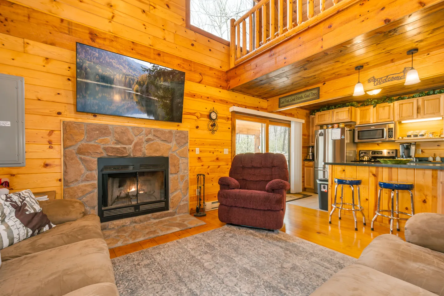 Living room with wood-paneled walls, stone fireplace, wall-mounted TV, and open view into the kitchen.