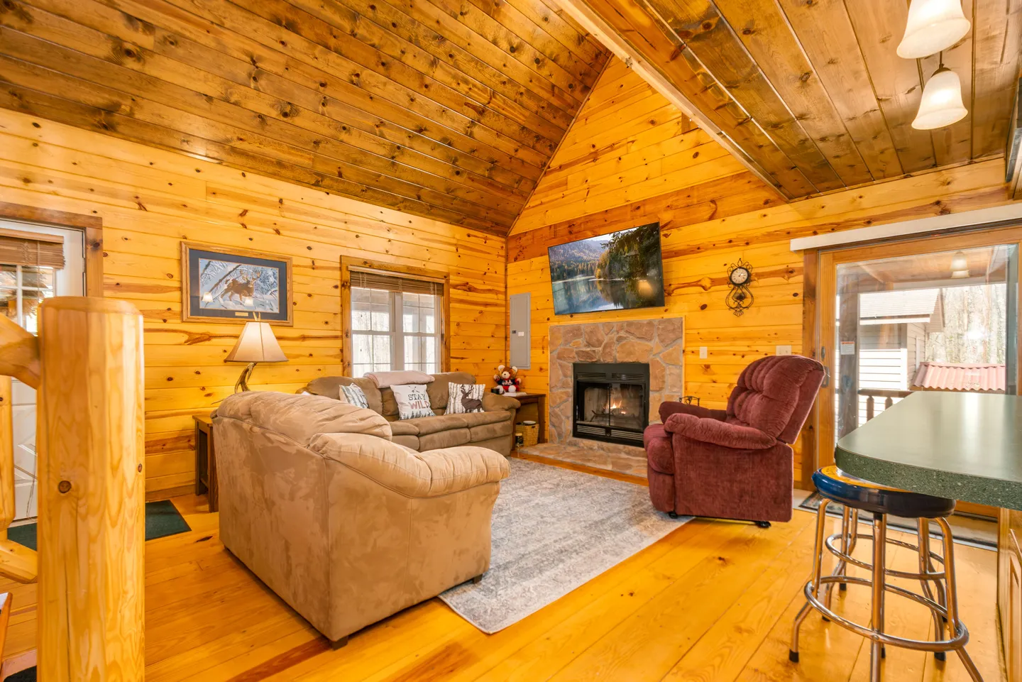 Living room with wood-paneled walls and ceiling, featuring sofas, an armchair, and a stone fireplace with a mounted TV.