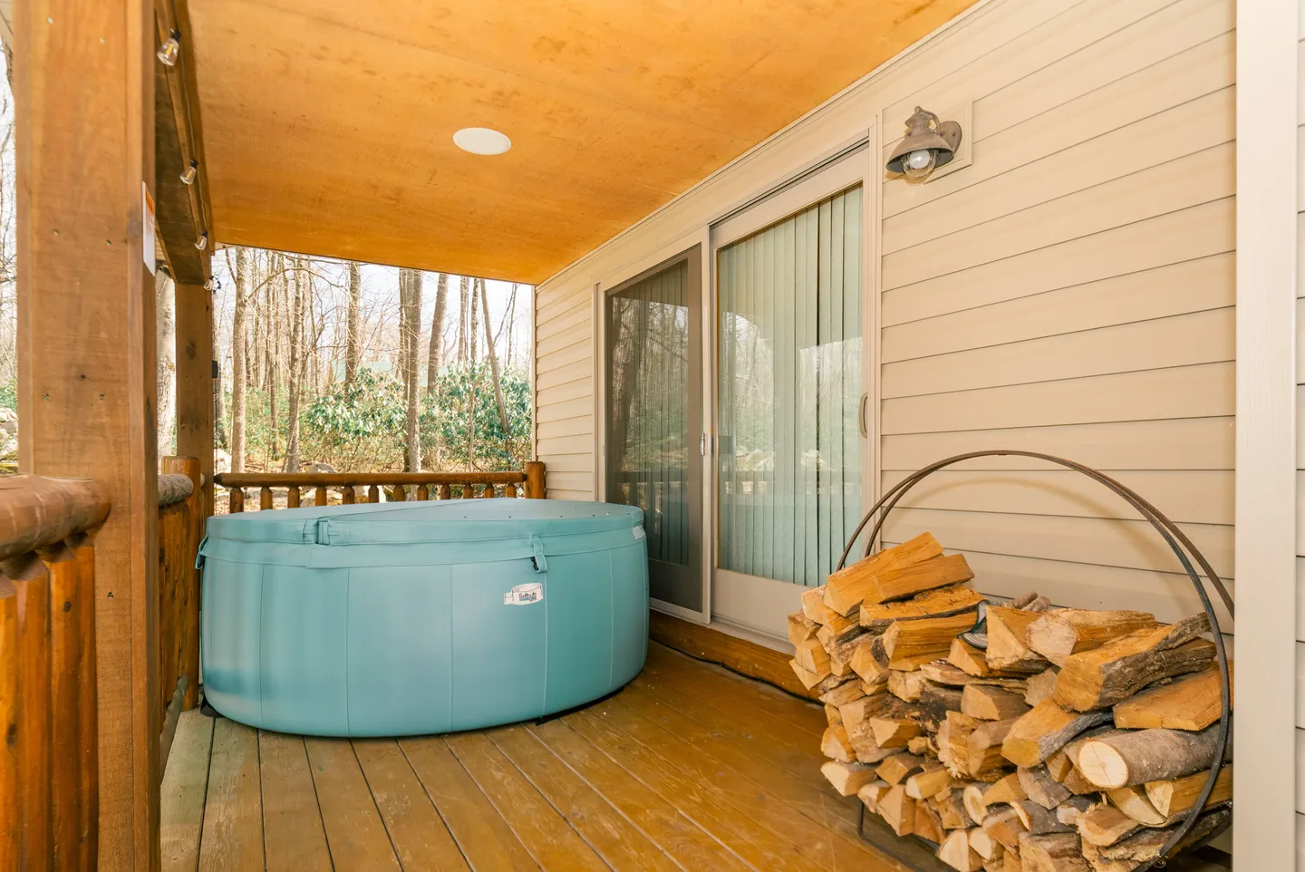 Covered wooden deck featuring a light blue hot tub, sliding glass patio doors, and a metal rack stacked with firewood.