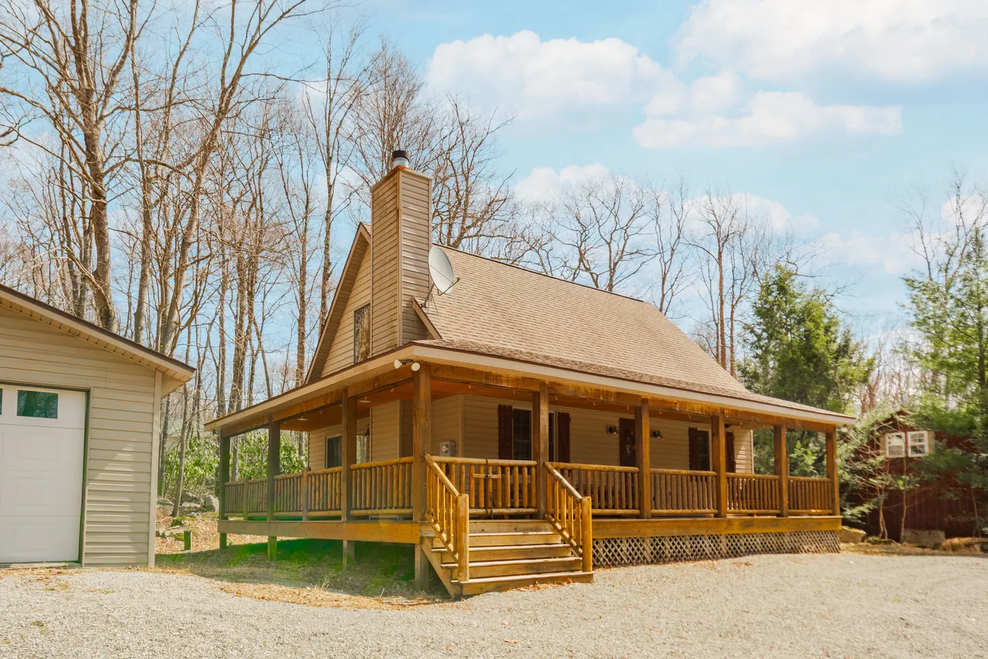 Exterior view of a wood-sided cabin with a wraparound covered porch, chimney, and gravel driveway next to a garage.