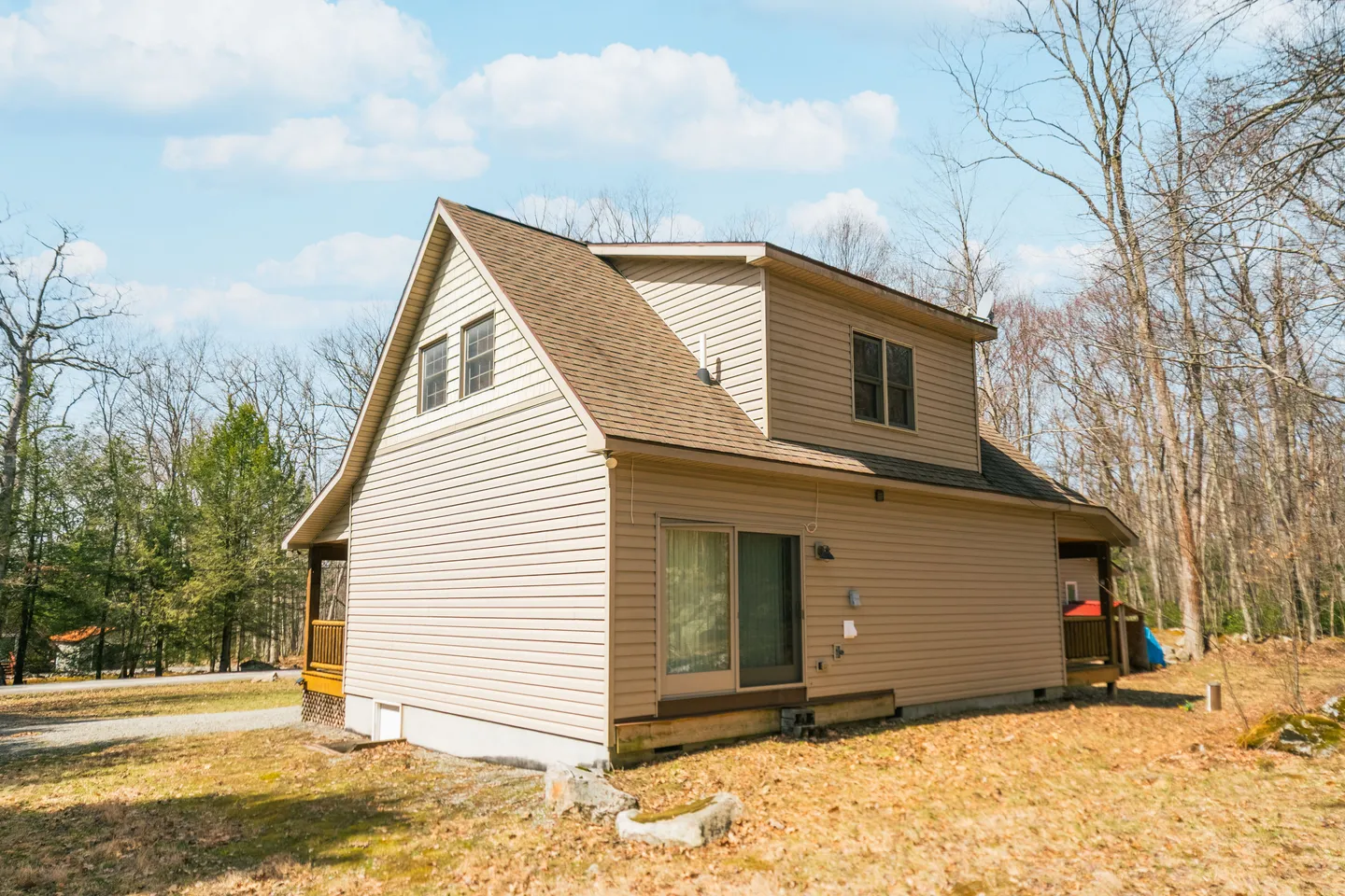 Exterior view of a two-story beige house with vinyl siding, shingled roof, sliding glass patio door, and a wooded yard under a blue sky.