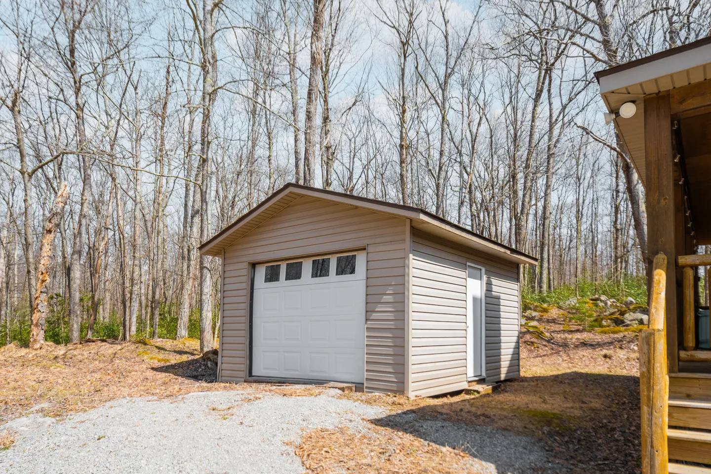 A detached single-car garage or storage shed with light beige siding, a white roll-up door, and a side entry door surrounded by trees.