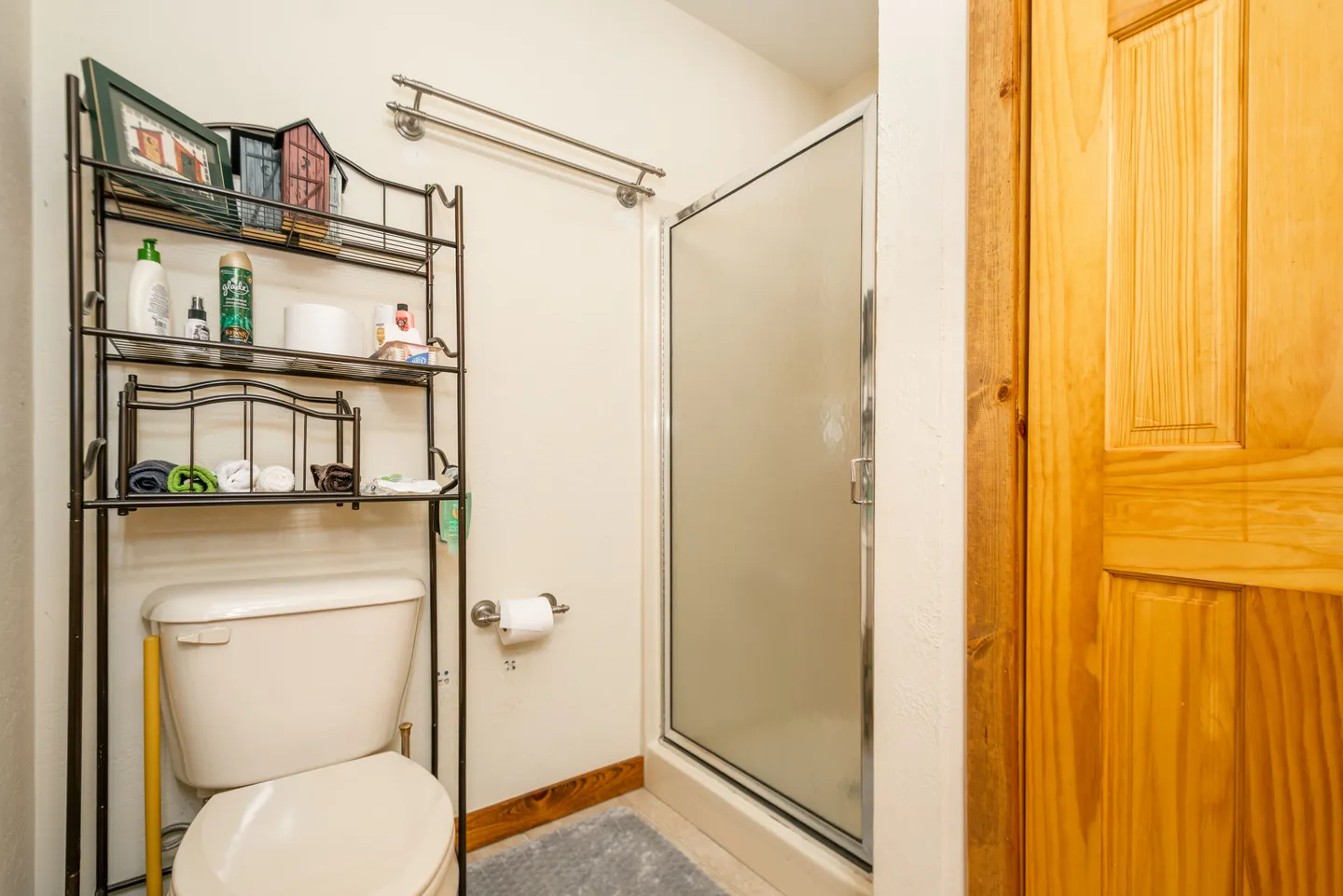 Bathroom showing a toilet beneath an over-the-toilet storage rack, a frosted glass shower door, and a wood door frame.