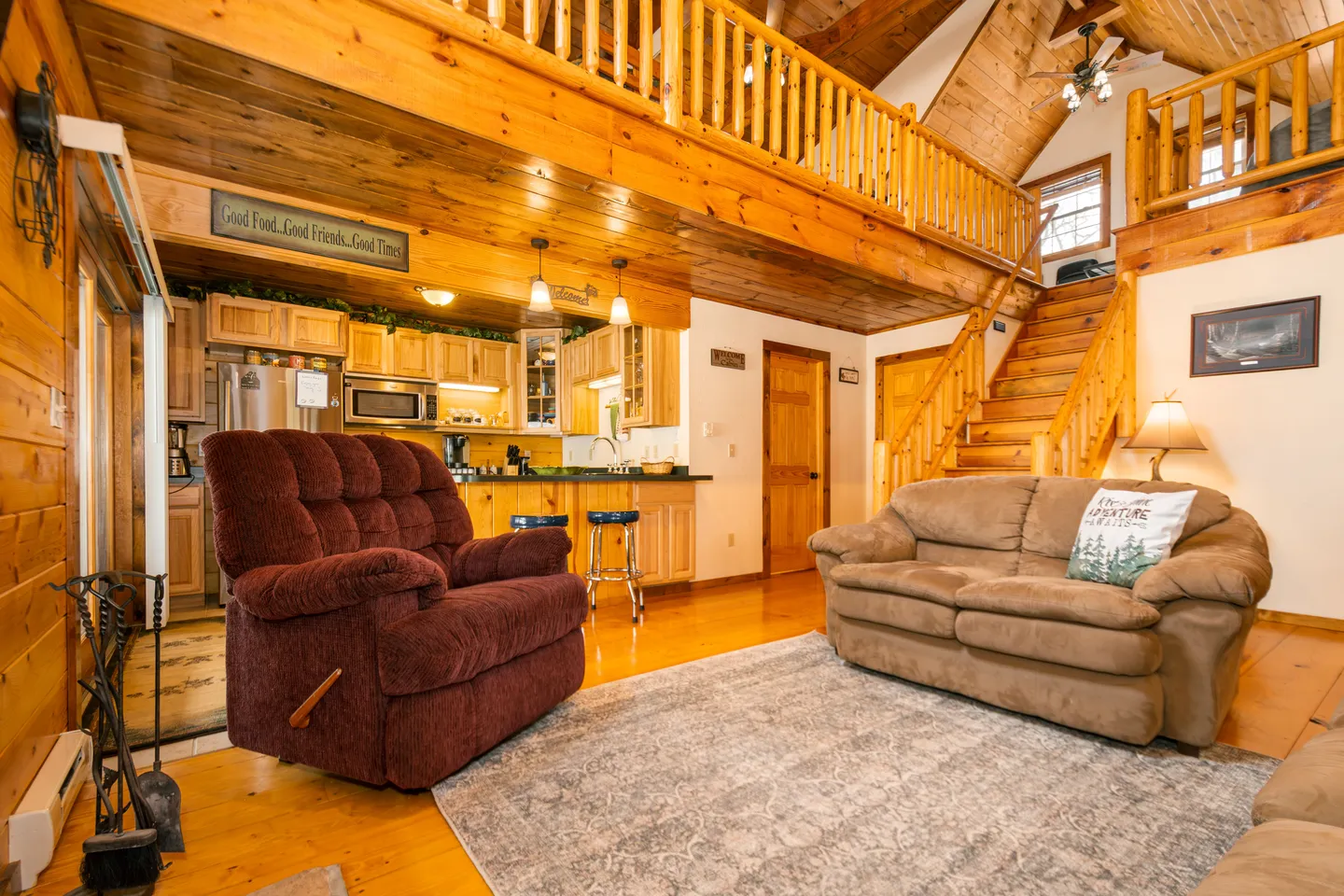 Open living area and kitchen inside a log cabin with wood-paneled walls, an open loft above, sofa, armchair, and rug.