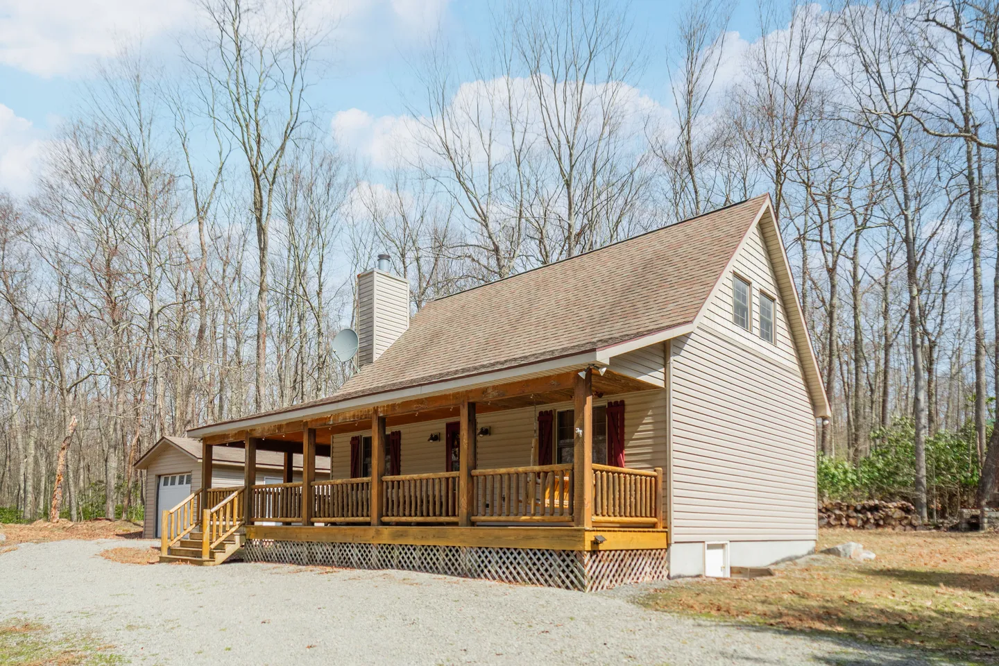 Exterior view of a cabin-style house with a covered front porch, gravel driveway, and detached garage surrounded by trees.