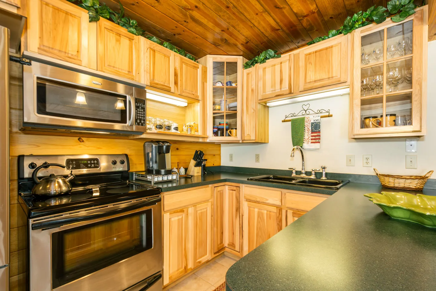Kitchen corner featuring light wood cabinetry, dark countertops, a double basin sink, and stainless steel stove with an over-the-range microwave.