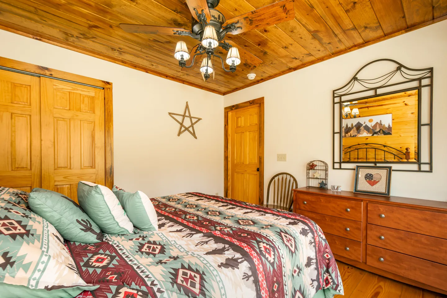 Bedroom with wooden plank ceiling, rustic ceiling fan, wood dresser with mirror, and bed dressed in patterned bedding.