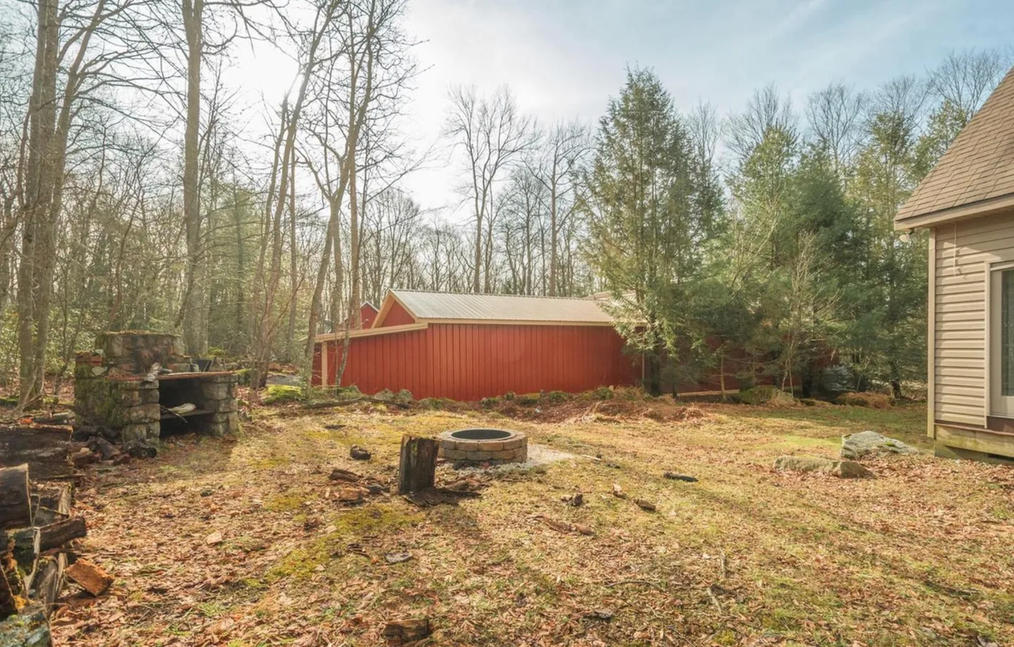 Wooded backyard with a stone outdoor fireplace, round stone fire pit, and red outbuilding in the distance.