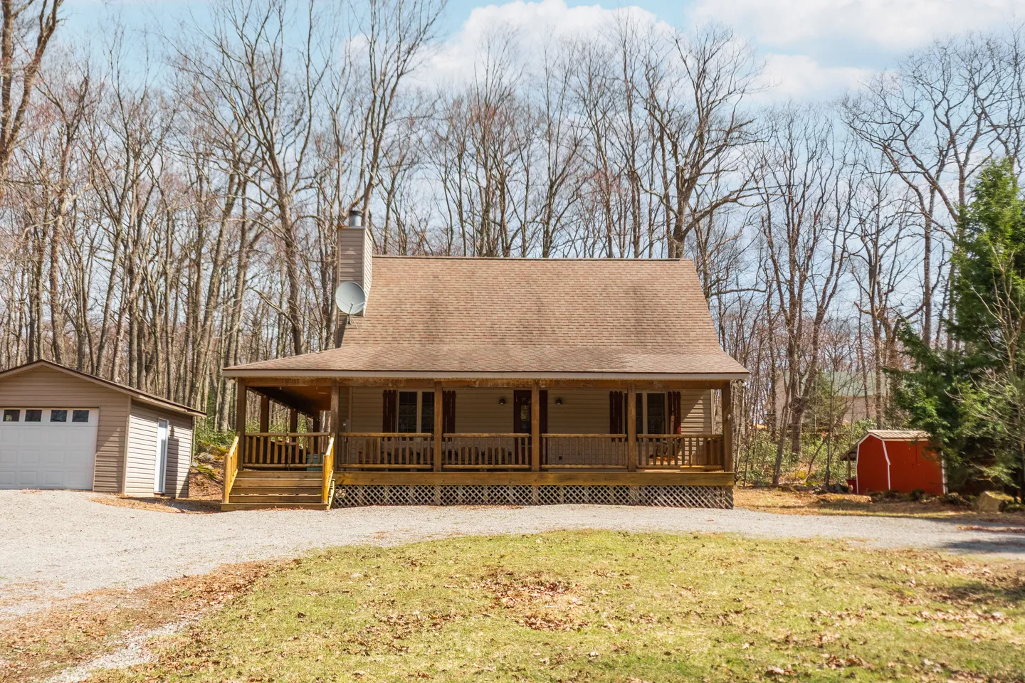 Exterior front view of a wood-sided cabin with a large covered front porch, surrounded by bare trees under a partly cloudy sky.