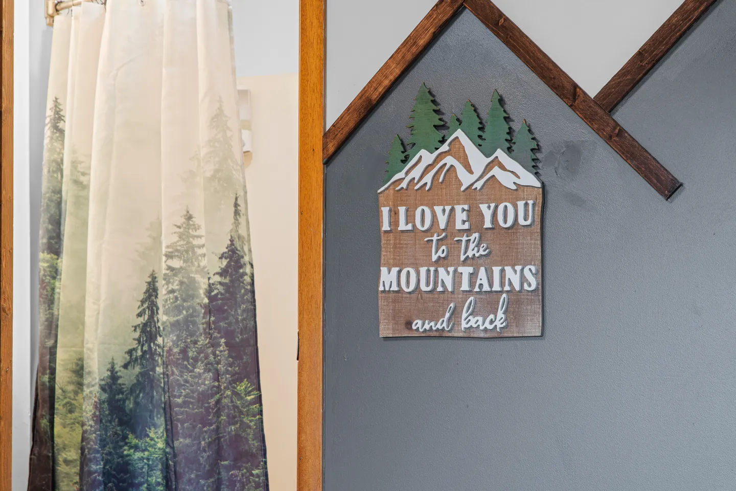 A close-up of a rustic wall sign reading 'I love you to the mountains and back' mounted on a gray wall next to a shower curtain with a forest print.