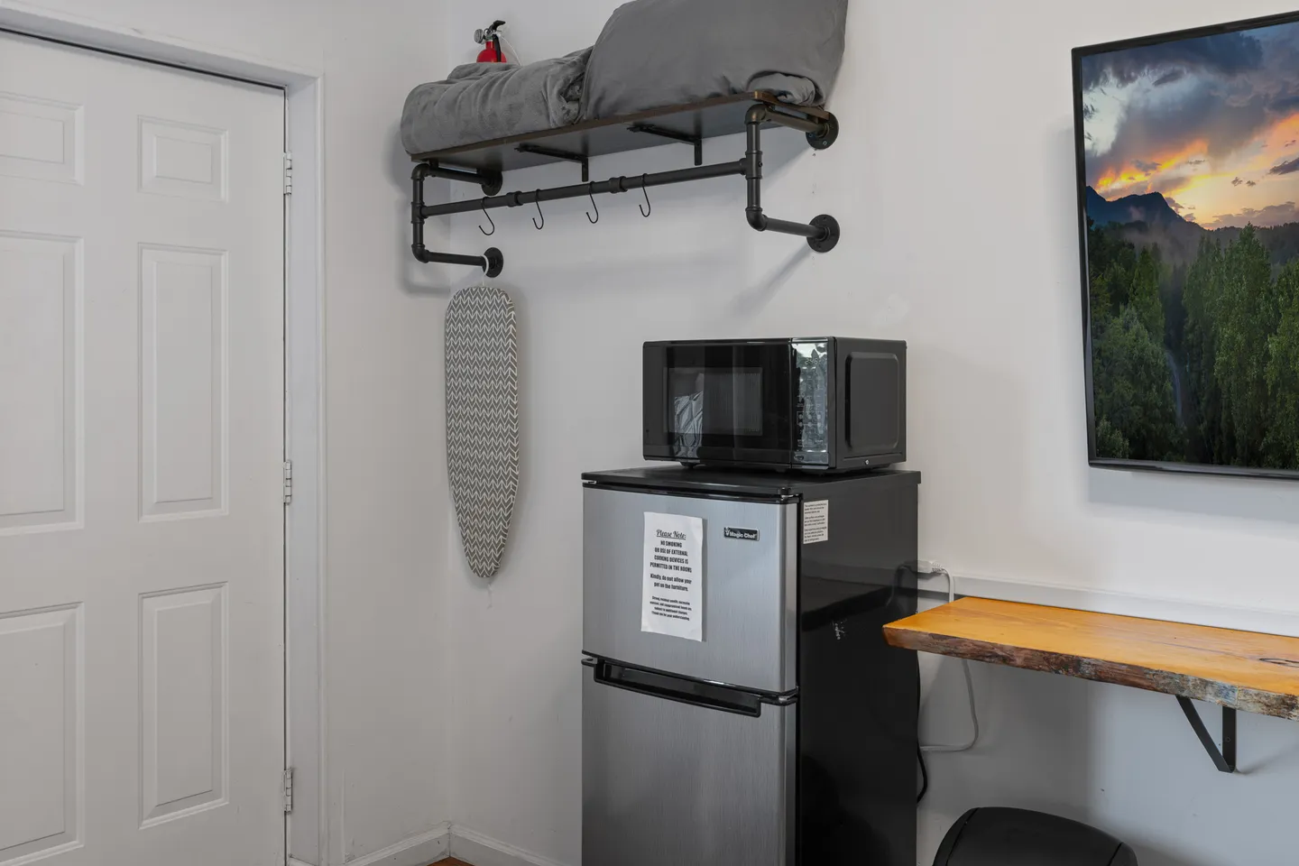 A corner of a room featuring a mini fridge, microwave, wall-mounted shelf with bedding, and an ironing board.