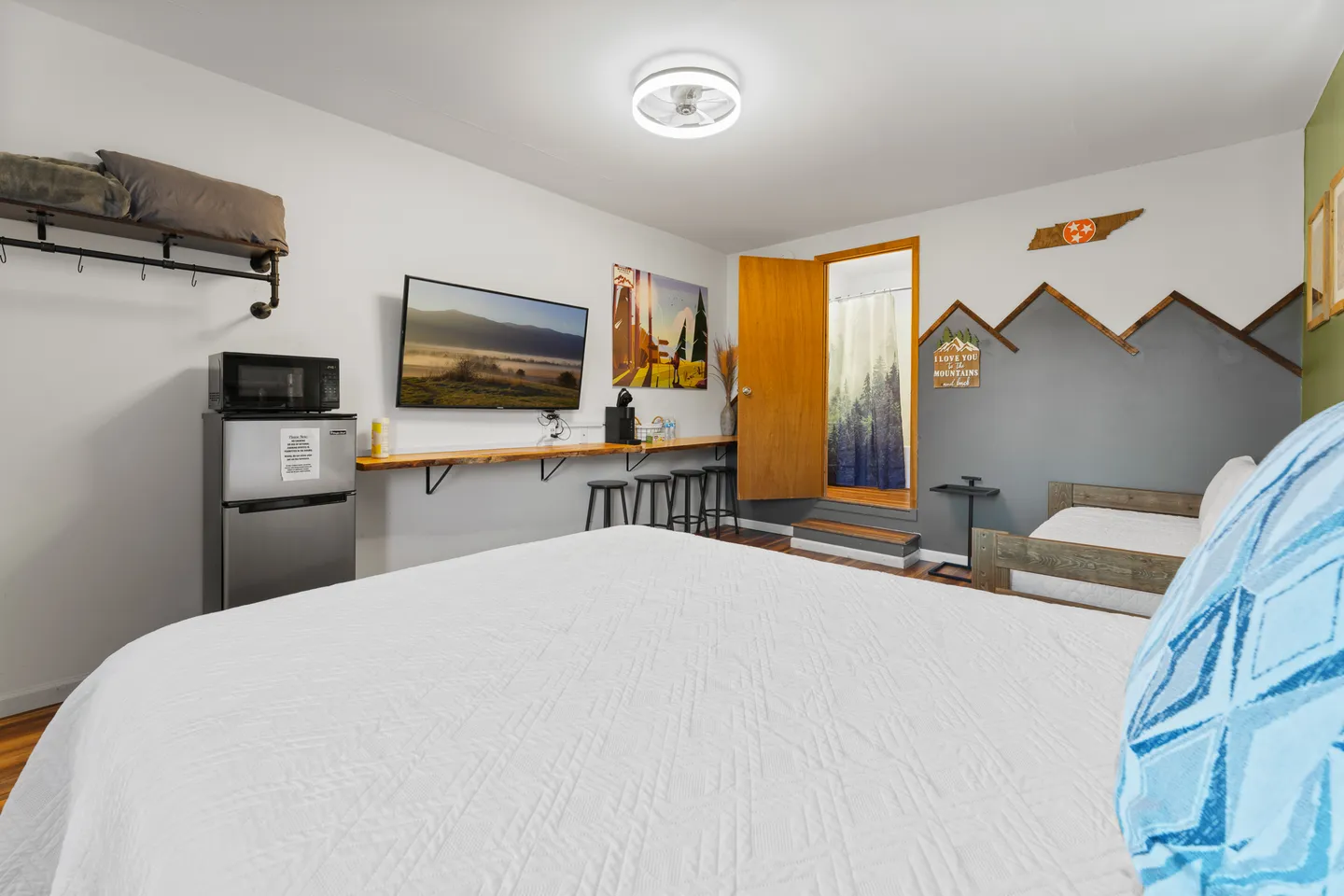 Bedroom featuring a bed with a white textured quilt, a wall-mounted TV, counter seating, mini fridge, and an open doorway.