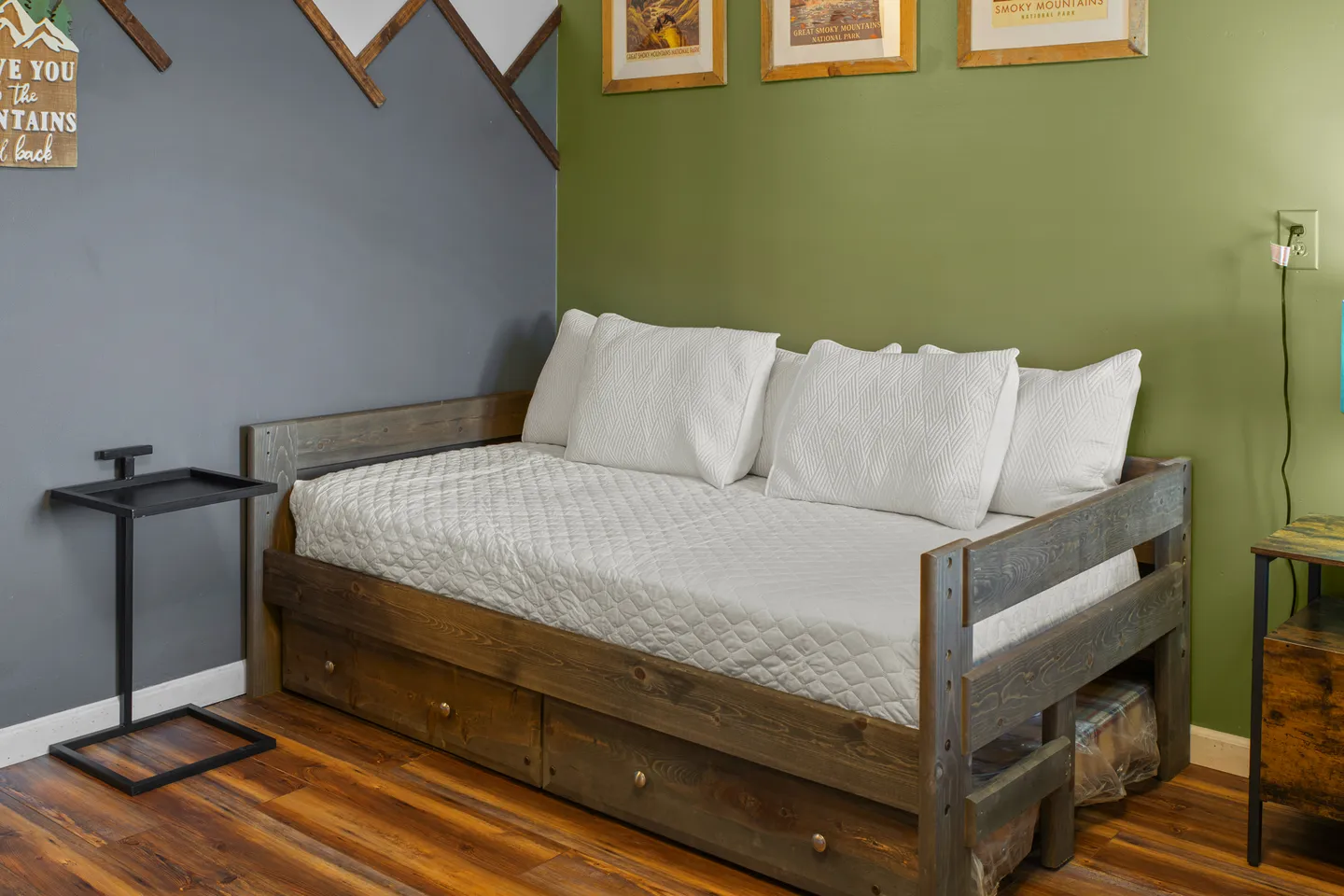 A wooden daybed with white quilted bedding and pillows set against a green and grey wall with wood-look flooring.