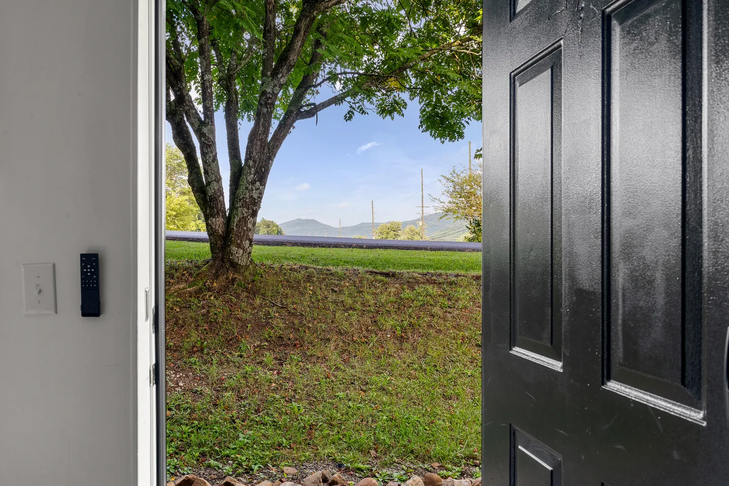 View looking out an open black front door toward a grassy slope, trees, a paved road, and distant mountain ridges.