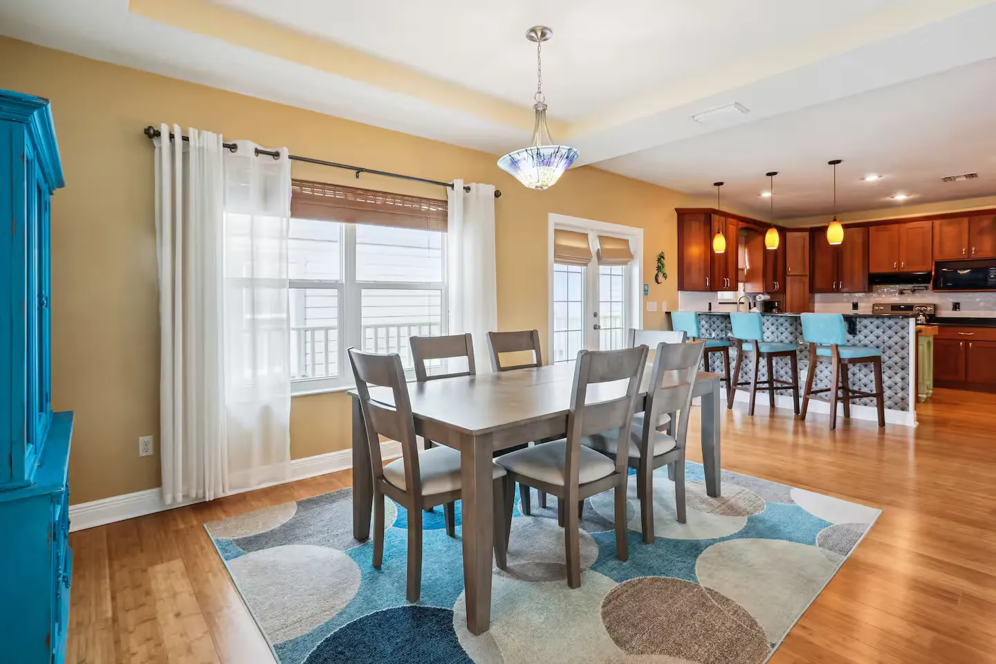 Dining area with a wooden table, chairs, patterned rug, and a kitchen with an island in the background.