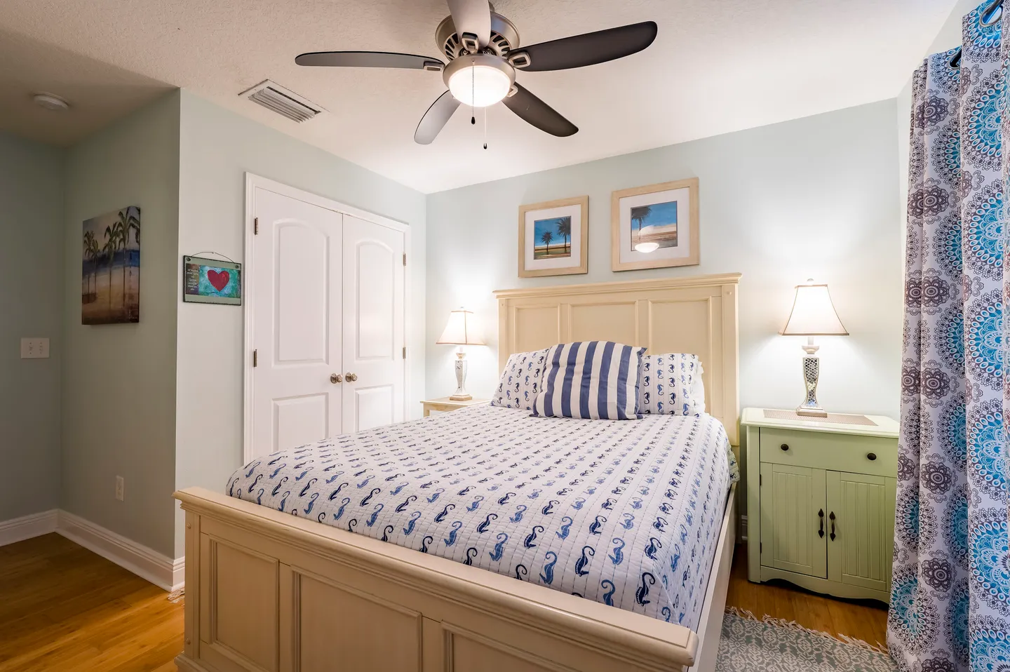 Bedroom with light blue walls, a cream-colored bed with patterned bedding, two nightstands with lamps, and a ceiling fan.