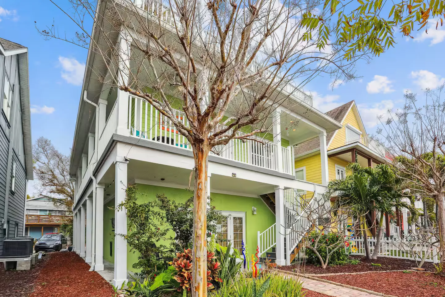 Exterior view of a multi-story light green vacation home with white support columns, covered porches, and landscaping.