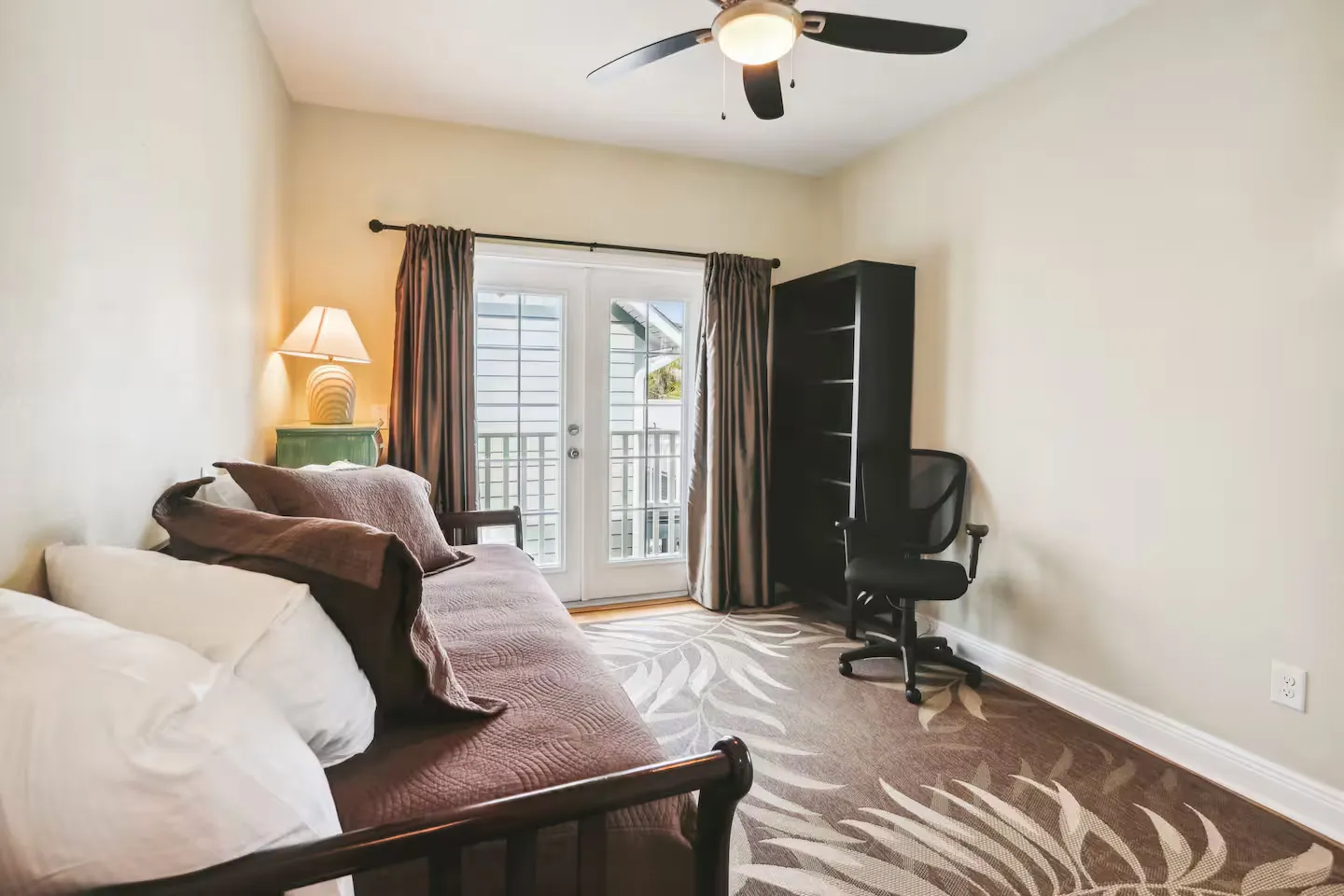Bedroom or office with a daybed, patterned rug, desk chair, bookcase, and French doors leading to an exterior balcony.
