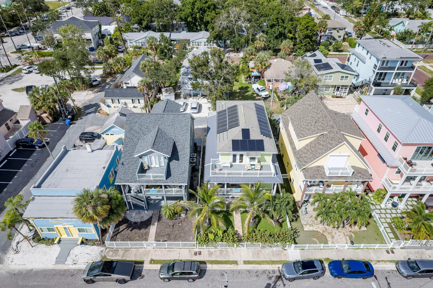High-angle aerial view of a row of coastal multi-story vacation homes along a paved residential street with parked cars and palm trees.