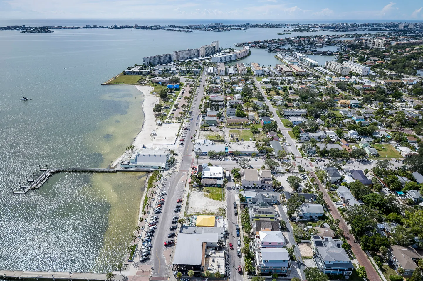 High aerial view of a coastal town featuring a beach park, long wooden pier, roadways, and surrounding residential neighborhoods next to a bay.