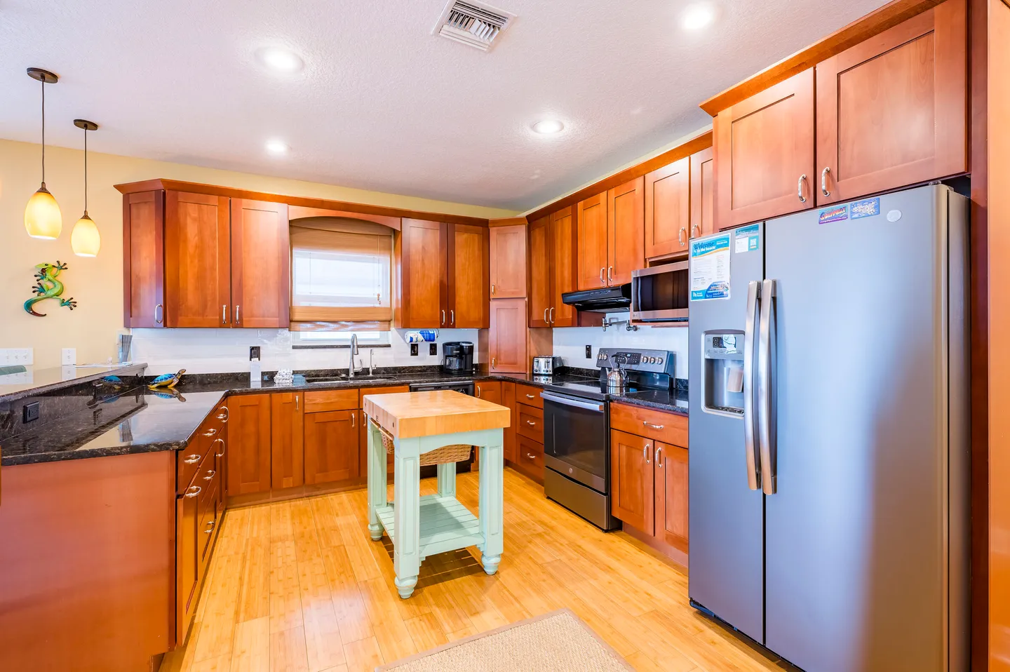 A kitchen with warm wood cabinetry, dark countertops, a stainless steel refrigerator, and a small light green mobile island.