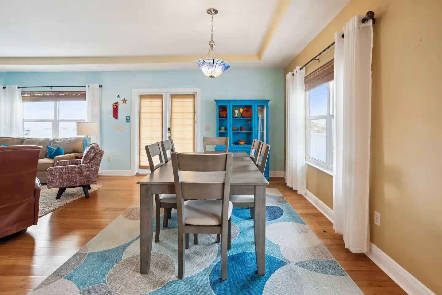 Dining area with a wooden table, grey chairs, patterned blue area rug, and an adjoining living room.
