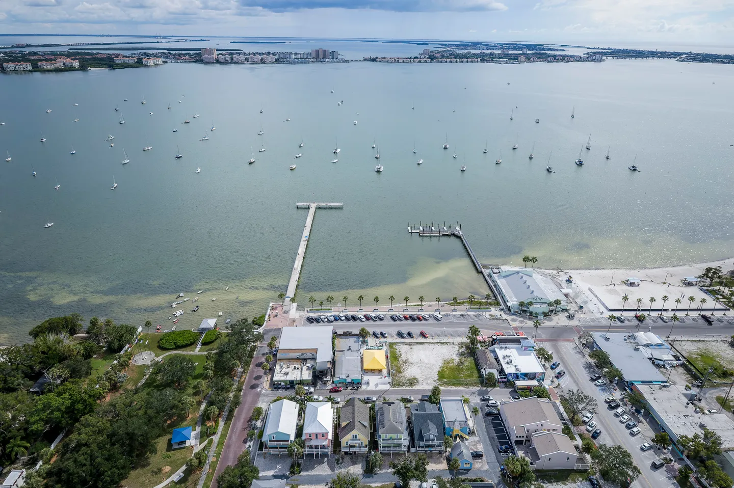 Aerial view of a coastal waterfront showing colorful houses, a long dock, and sailboats anchored in a bay.