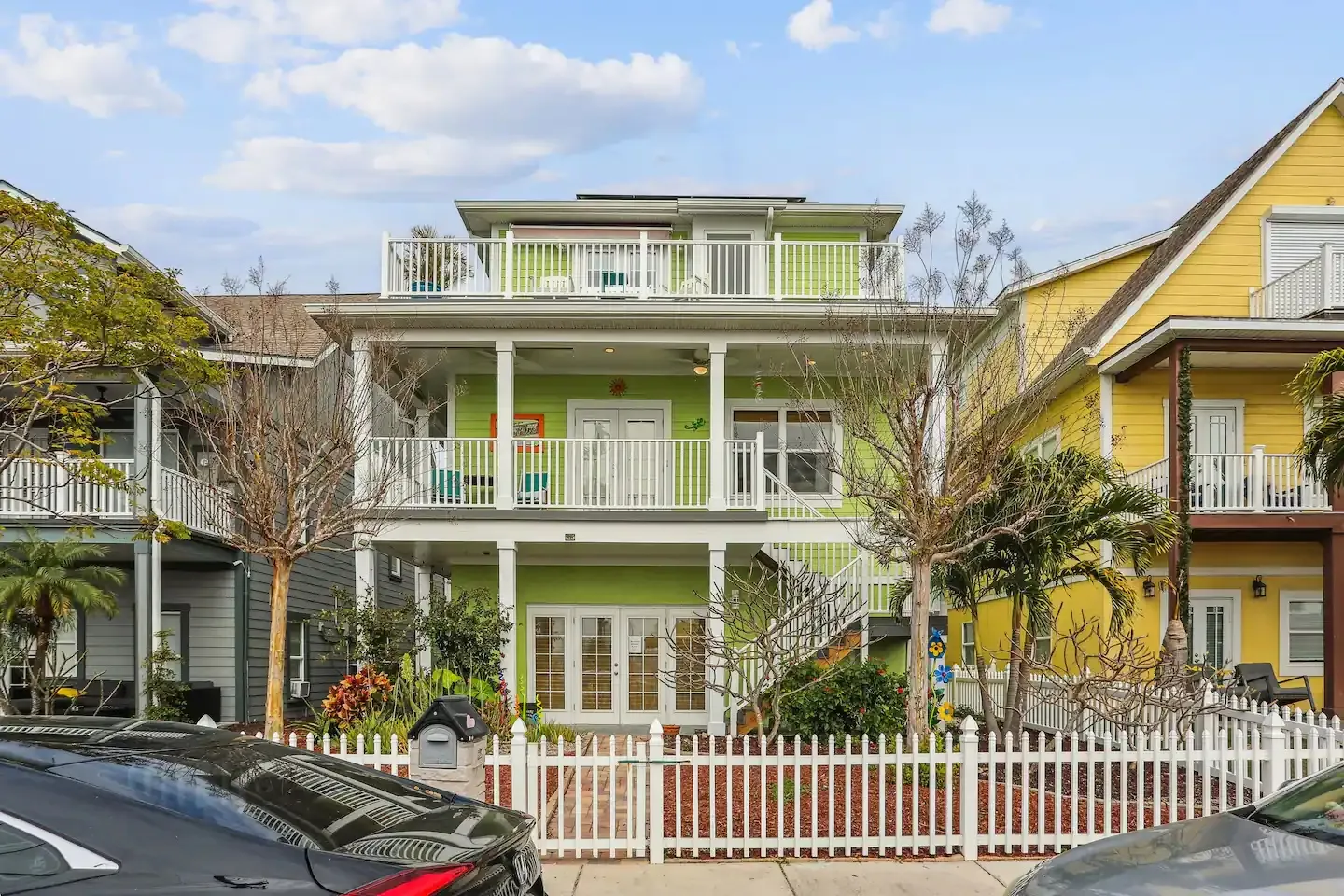 Exterior front view of a multi-story light green beach home with white balconies, stairs, and a white picket fence.