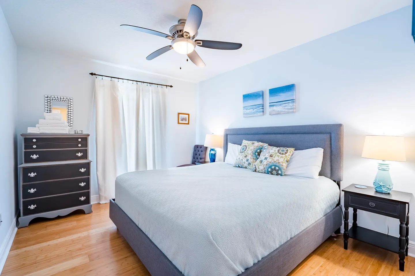 Bedroom with an upholstered gray bed, wood floors, dark nightstand and dresser, and coastal artwork on light blue walls.