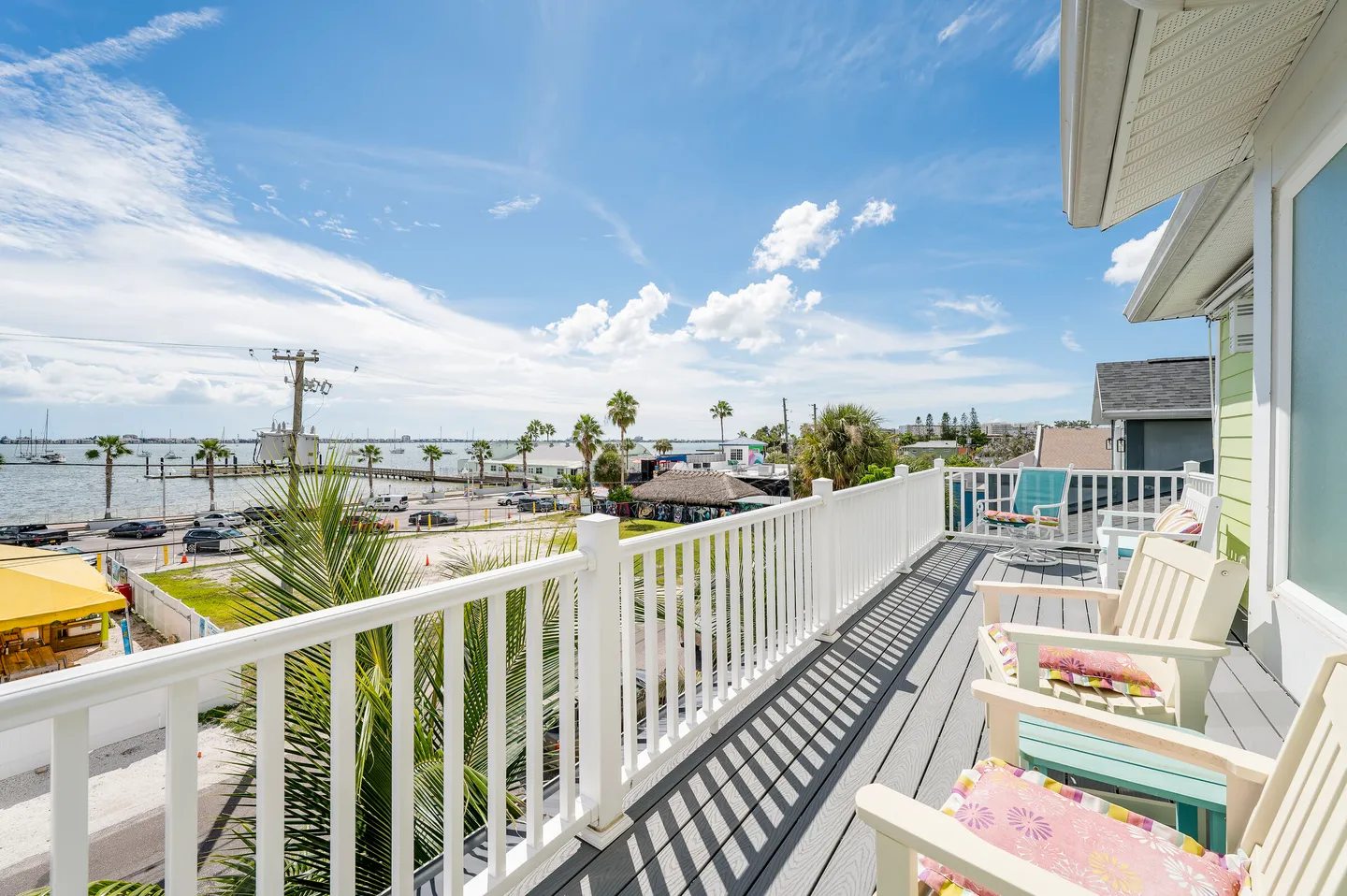 Elevated outdoor balcony with white railings, deck seating, and views of palm trees, water, and parked boats under a bright blue sky.