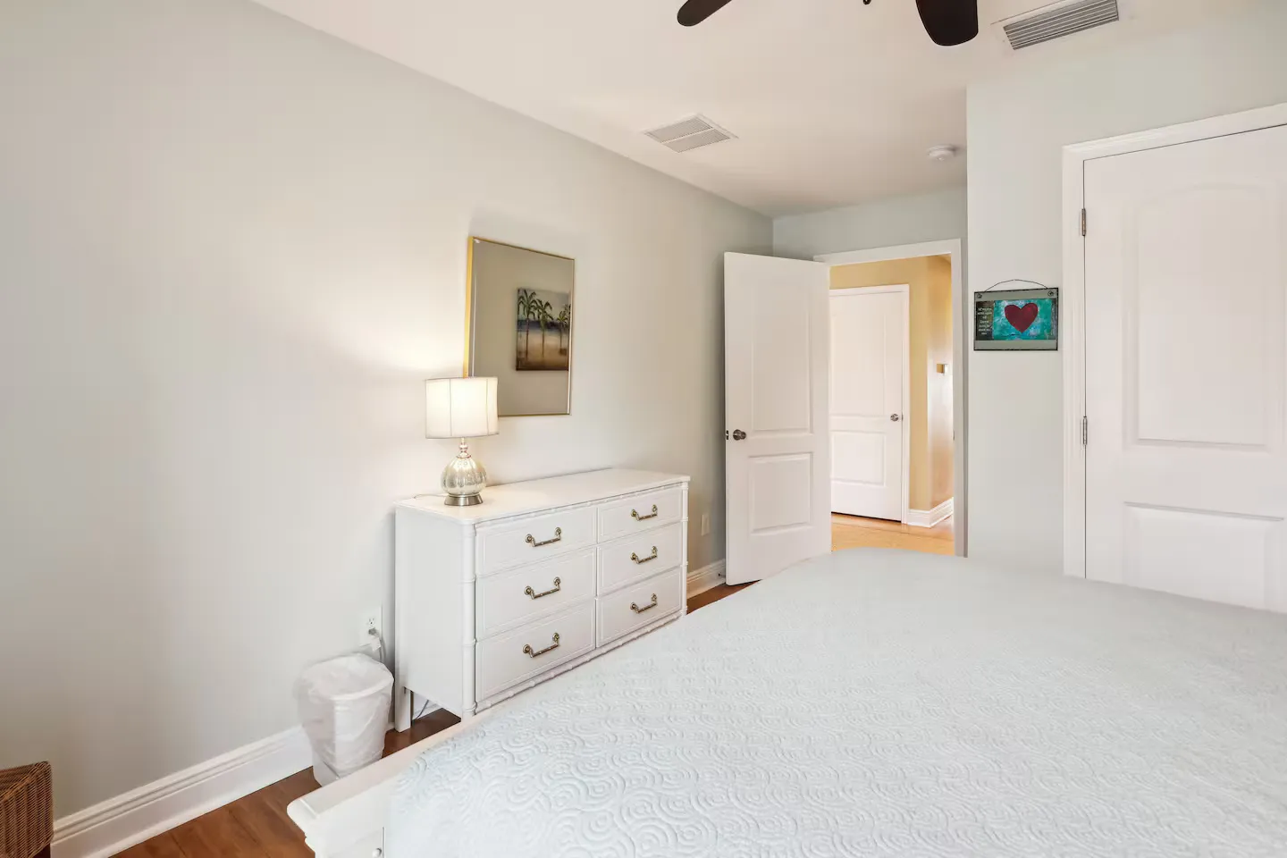 Bedroom featuring a made bed with a light-colored patterned coverlet, a white dresser with a lamp and mirror, and an open doorway leading to a hallway.