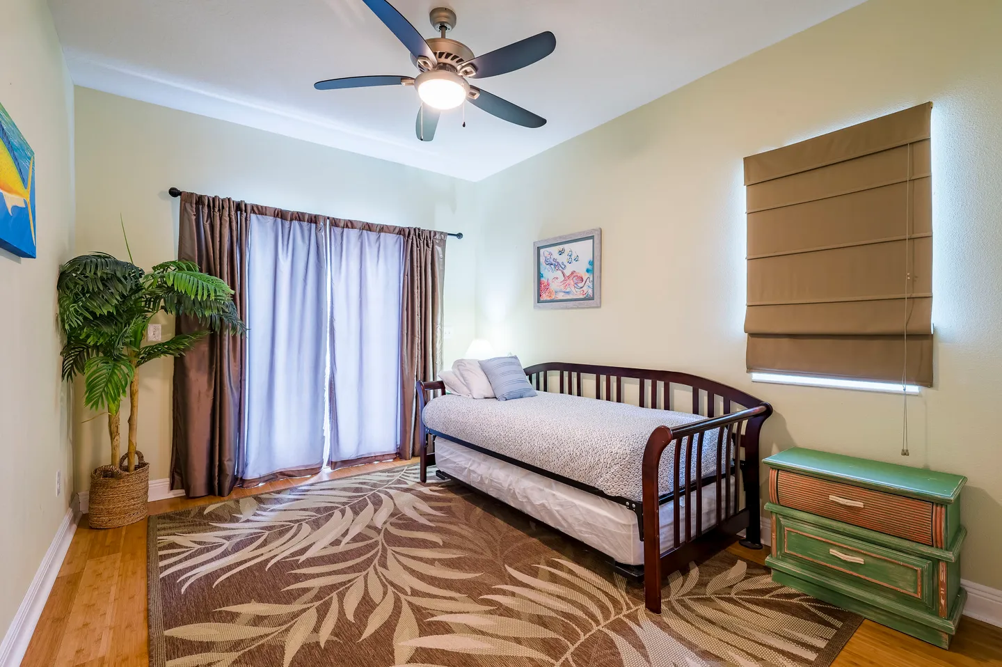 Bedroom with a wooden daybed and trundle, ceiling fan, brown curtains over sliding glass doors, and a leaf-patterned area rug.