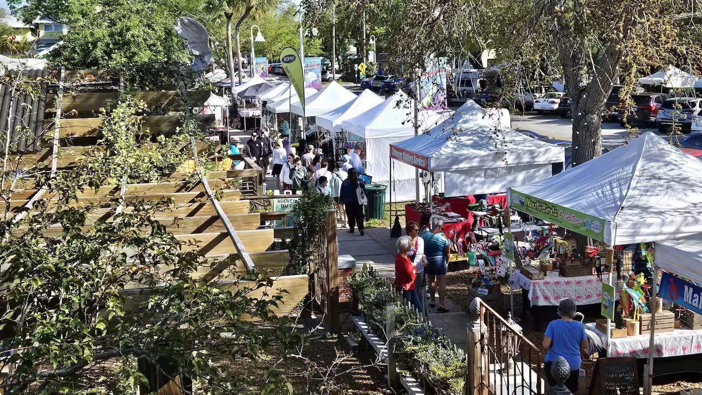 Elevated view of an outdoor street market with white vendor tents, trees, and browsing visitors under bright sunlight.