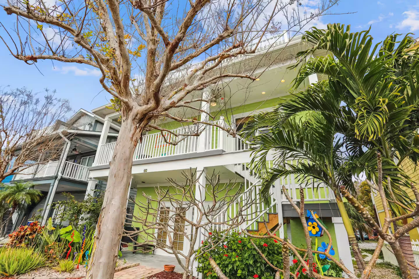 Low-angle exterior view of a multi-story light green home with white railings, framed by palm trees and landscaping.