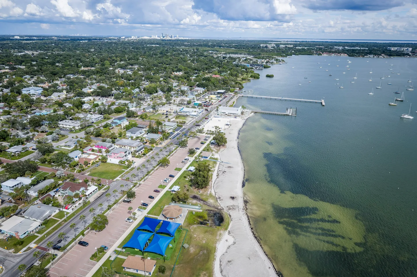 Aerial view of a coastal town featuring a sandy shoreline, piers extending into the bay with moored boats, parking areas, and residential neighborhoods.