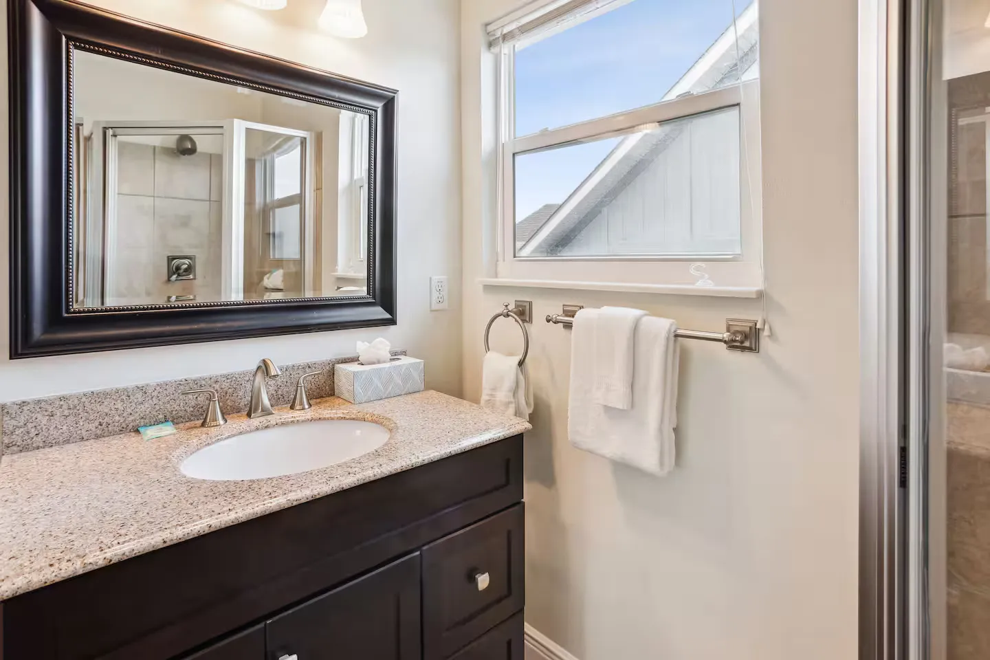 Bathroom vanity with dark cabinets, speckled countertop, and large framed mirror next to a window and shower.