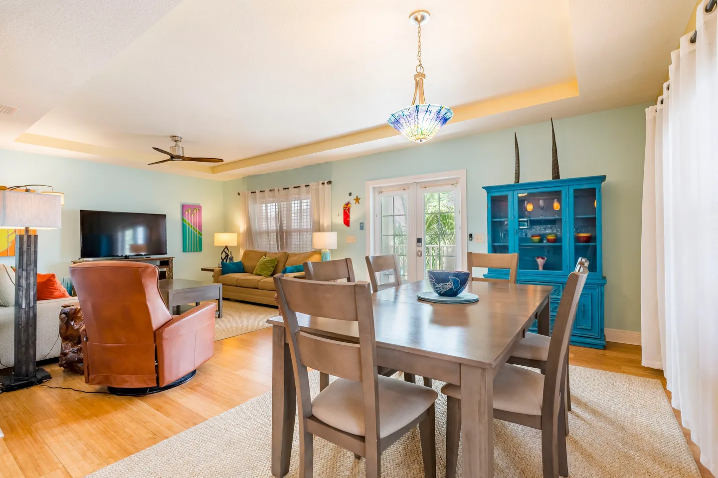 Open-concept dining and living room with wood dining table, tray ceiling, vibrant blue hutch, and living area with sofas and TV.