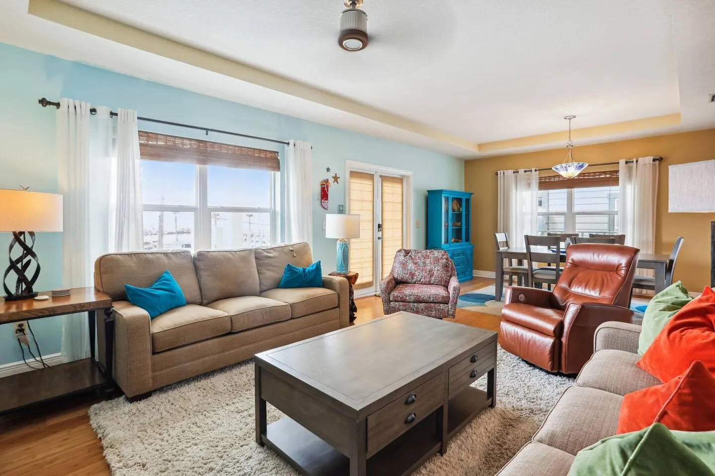 Living room with light blue walls, tan sofas, a coffee table, leather recliner, and an adjacent dining area in the background.