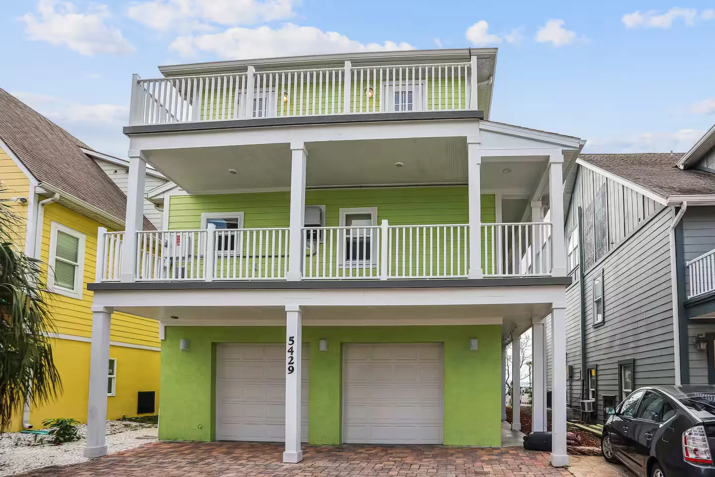 Exterior front view of a multi-story lime green coastal house with two garage doors and multiple white-railed balconies.