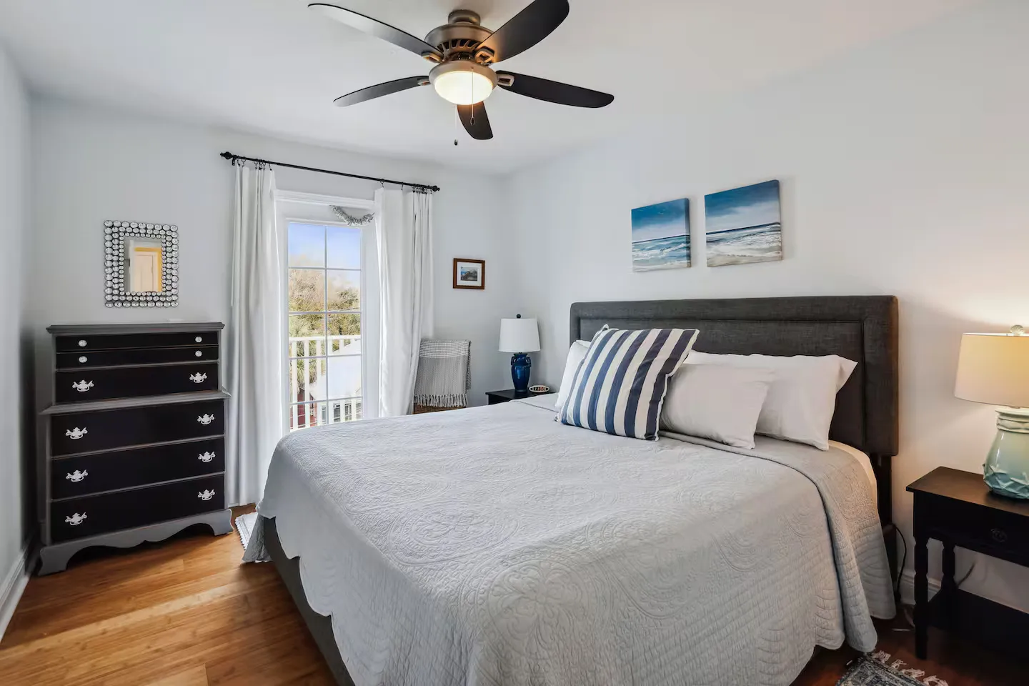 Bedroom with a large bed, ceiling fan, dark nightstands with lamps, a dresser, and glass doors leading to a balcony.