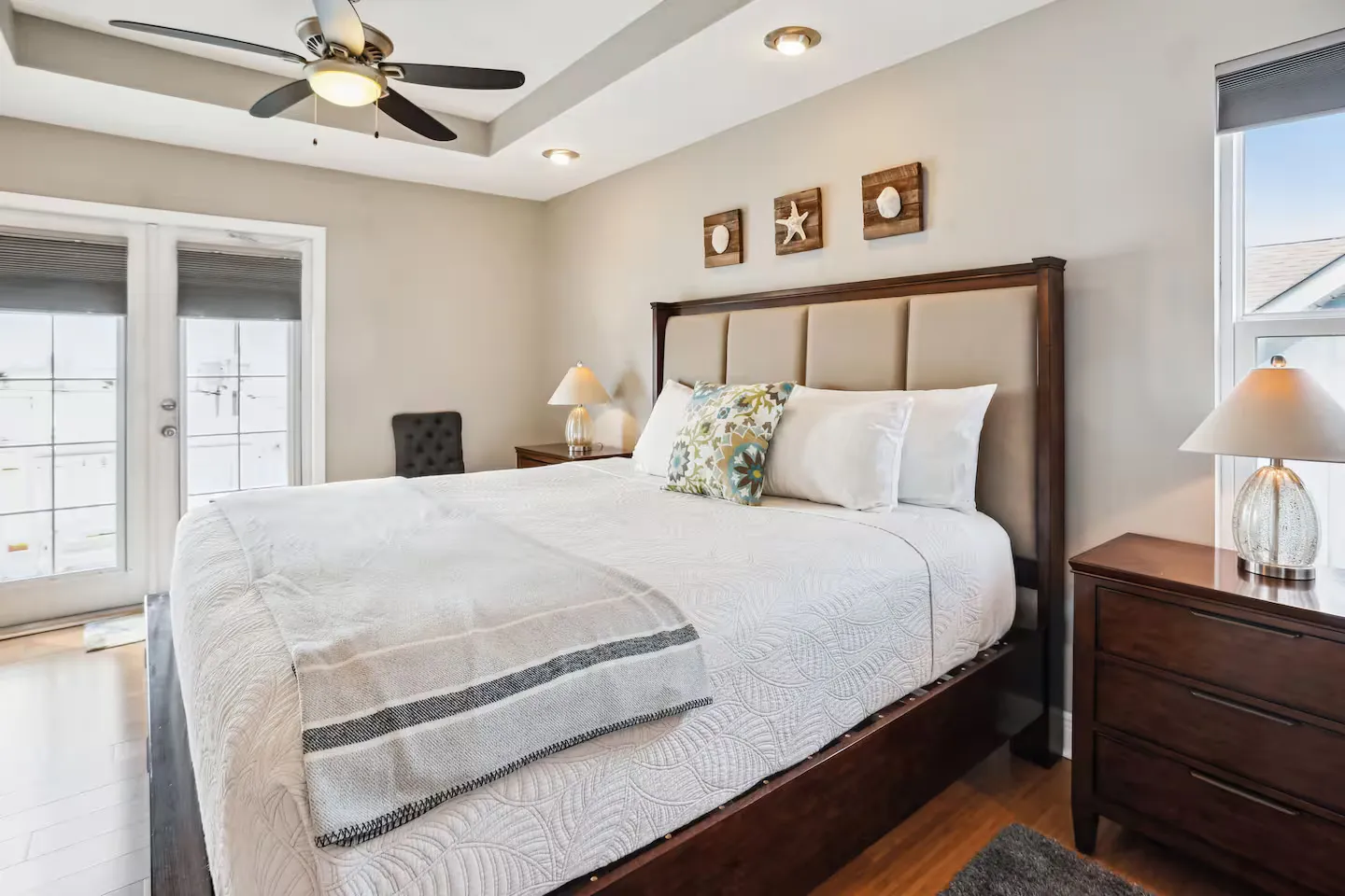 Bedroom featuring a made bed with an upholstered headboard, flanked by nightstands with lamps, tray ceiling with fan, and glass doors leading outside.