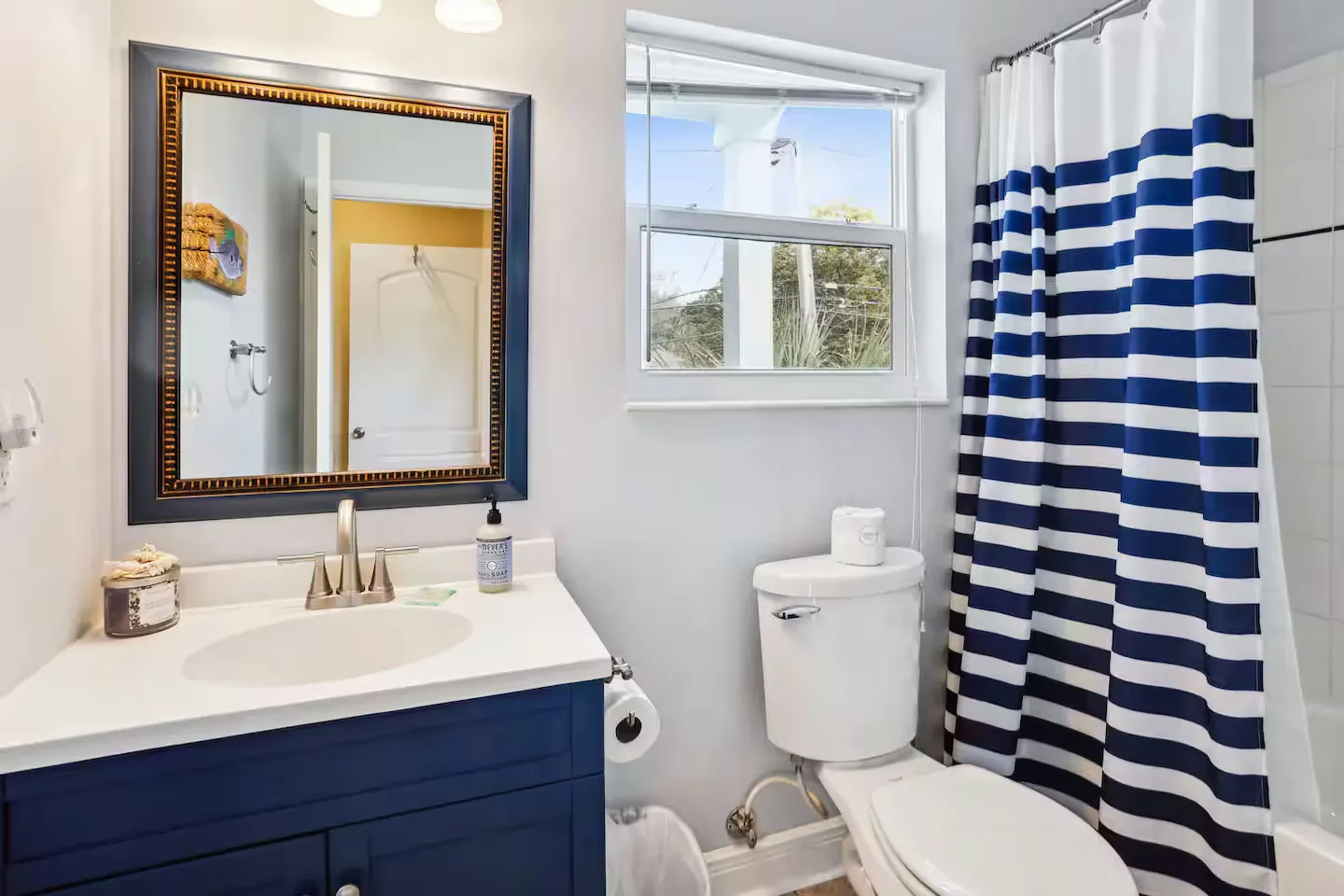 Bathroom with a blue vanity, white countertop, framed mirror, toilet, window, and blue and white striped shower curtain.