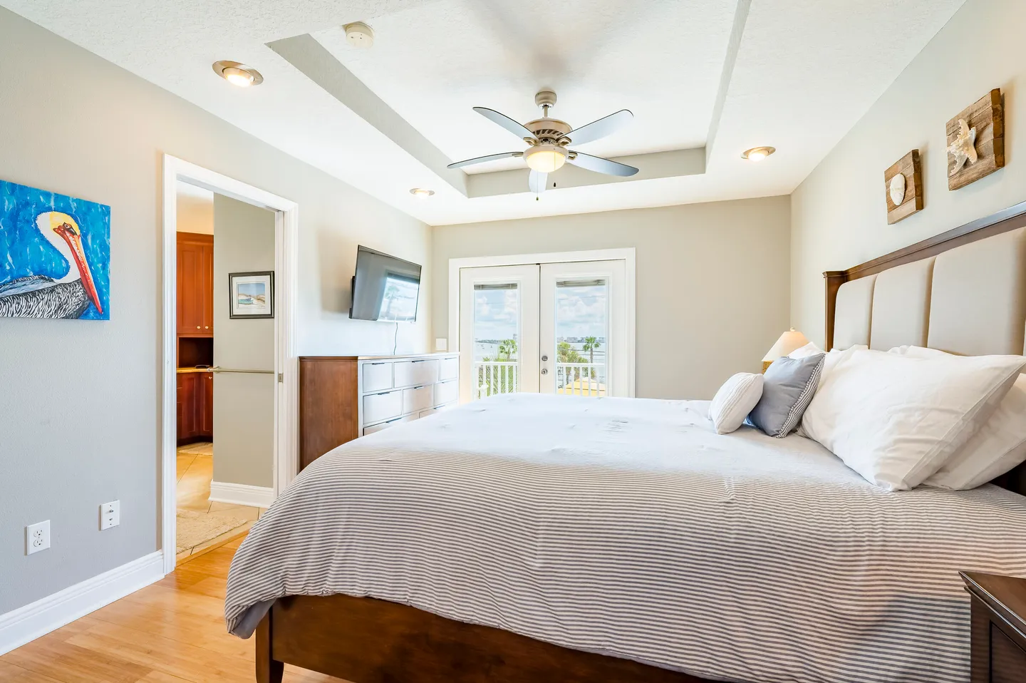Bedroom with a large bed, ceiling fan in a tray ceiling, dresser with a wall-mounted TV, and glass doors opening to a balcony with water views.