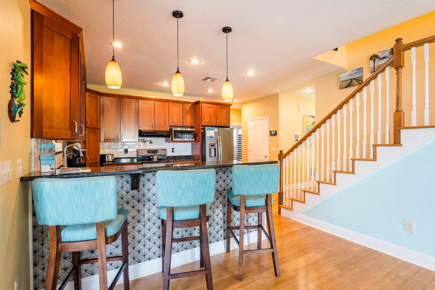 Kitchen counter with three turquoise barstools next to a staircase with white spindles and wooden railing.