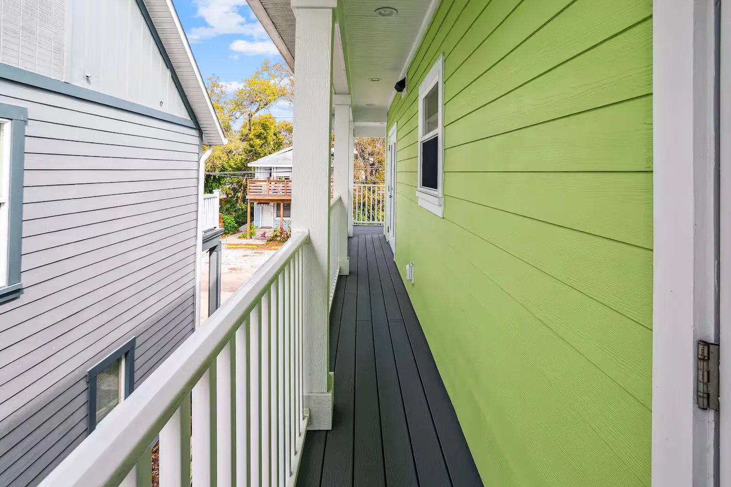 Narrow covered outdoor balcony with dark plank flooring, white railings, and bright green lap siding.