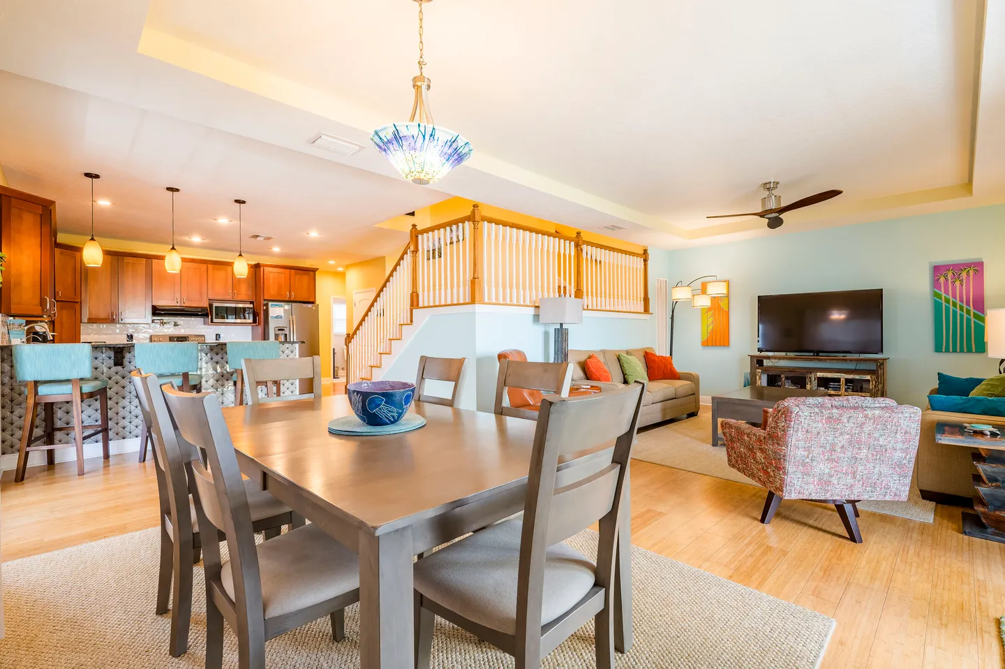 Open-concept living space with a wooden dining table and chairs in the foreground, adjacent to a kitchen with bar seating and a living area with a television.